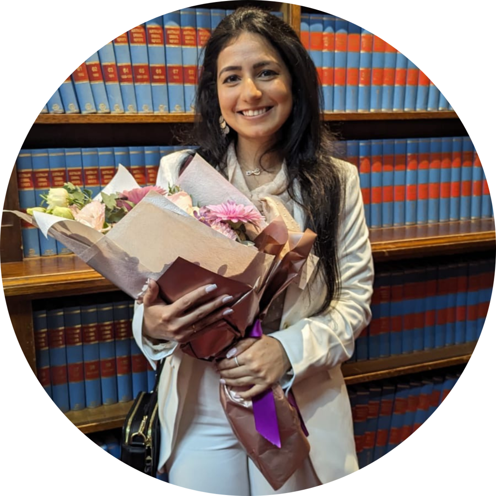 Ananya is holding a big bunch of flowers and looking directly at the camera. She is standing in front of a big bookshelf of legal books.