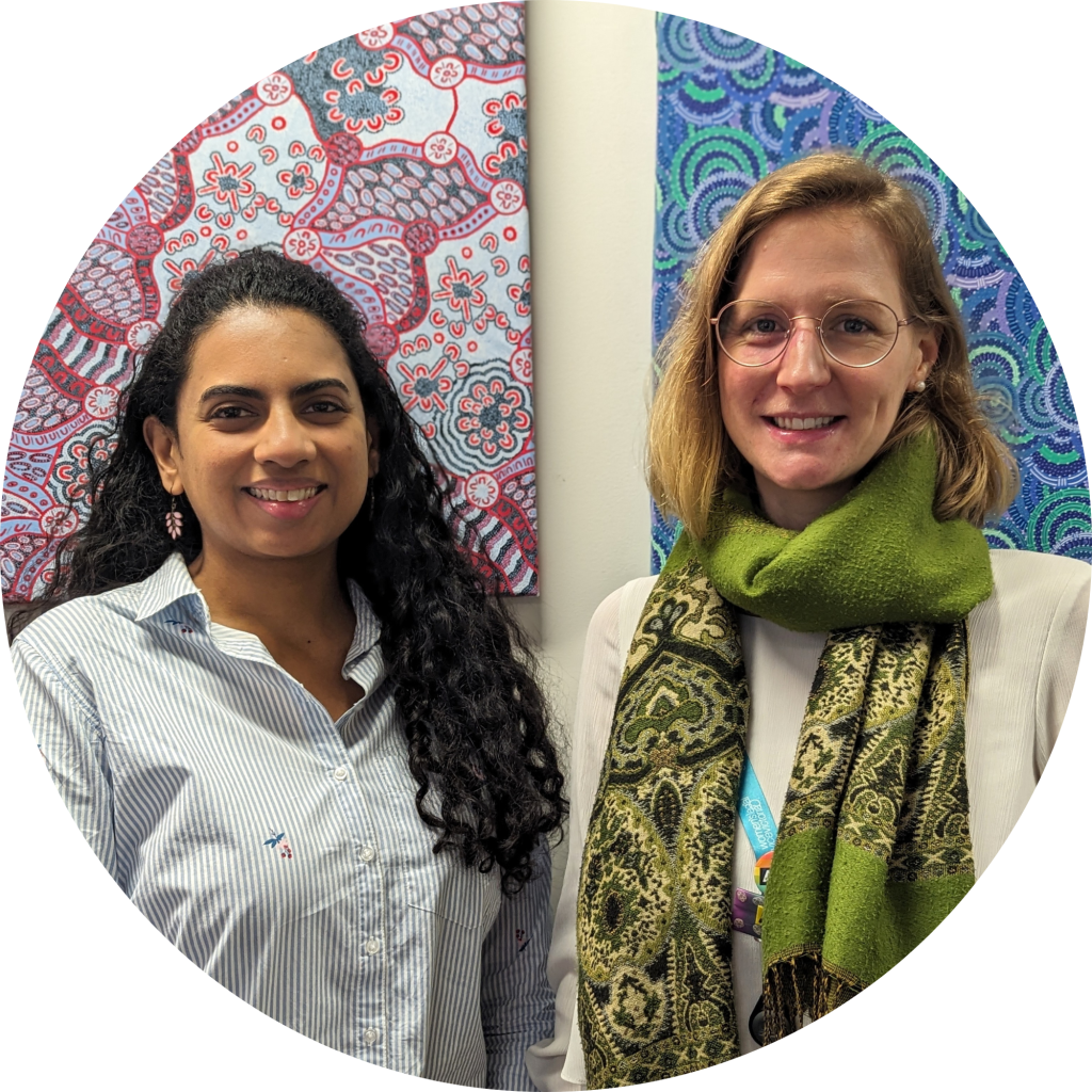 Dinesh and Anna are standing next to each other looking at the camera and smiling. They are standing in front of two Indigenous artworks.